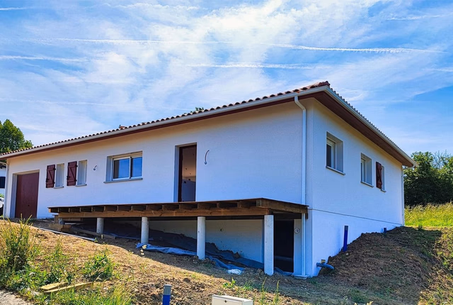 Façade de maison de plain-pied blanche avec terrasse en bois sur pilotis et volets bordeaux.