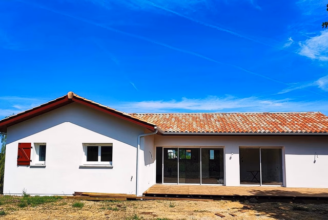 Façade blanche d'une maison de plain-pied avec toiture en tuiles, grandes baies vitrées et terrasse en bois.