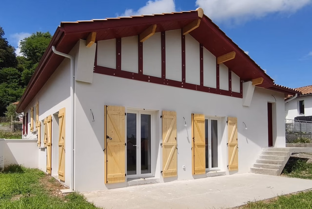 Façade blanche de plain-pied avec colombages rouges, volets en bois clair et terrasse en béton.