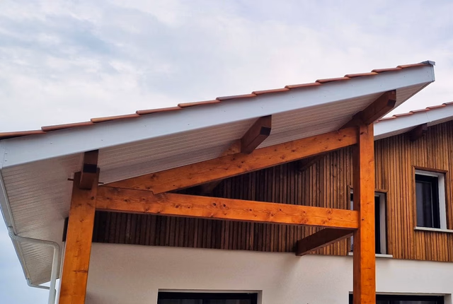 Avancée de toit soutenue par une charpente en bois massif devant une façade claire et bardée.