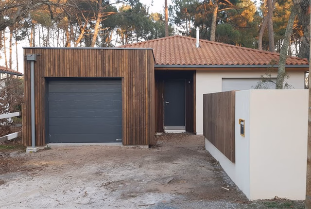 Façade d'une maison de plain-pied avec garage en bois, porte grise et muret d'entrée en crépi blanc.