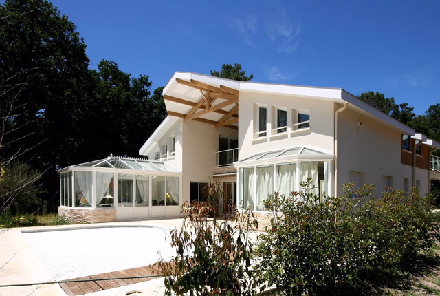 Grande maison à étage aux murs blancs avec deux vérandas vitrées entourant une piscine extérieure.