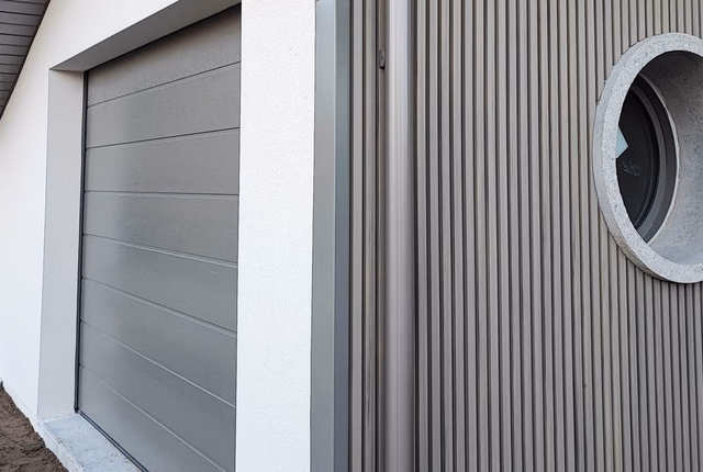Façade de maison avec porte de garage grise, bardage vertical et hublot rond encadré de pierre.