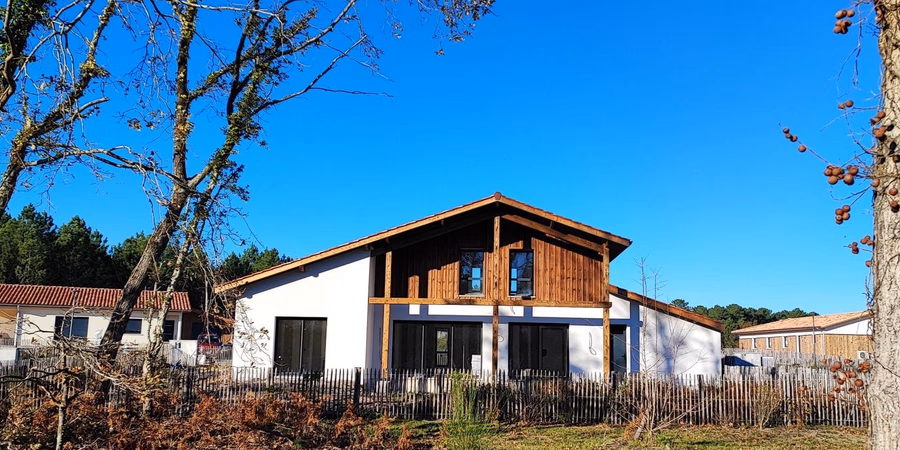 Villa contemporaine avec murs blancs et bardage en bois, entourée d'arbres sous un ciel bleu.