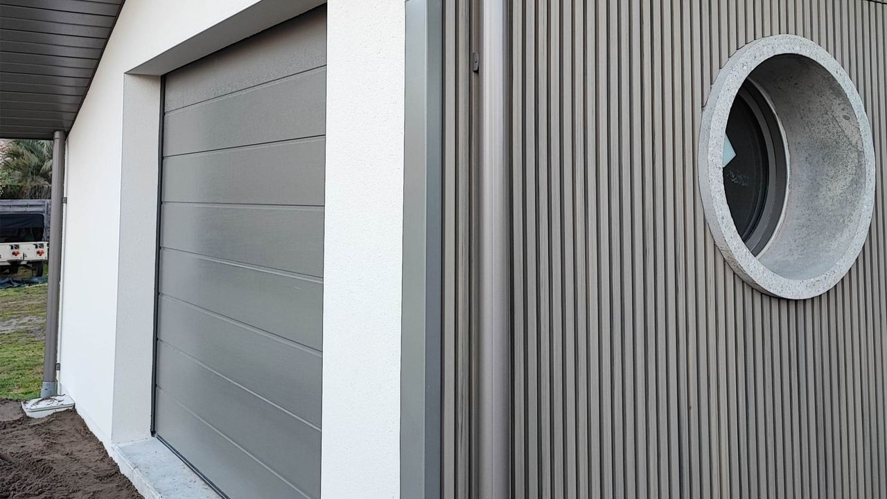 Porte de garage grise avec un mur en panneaux verticaux et une fenêtre circulaire.