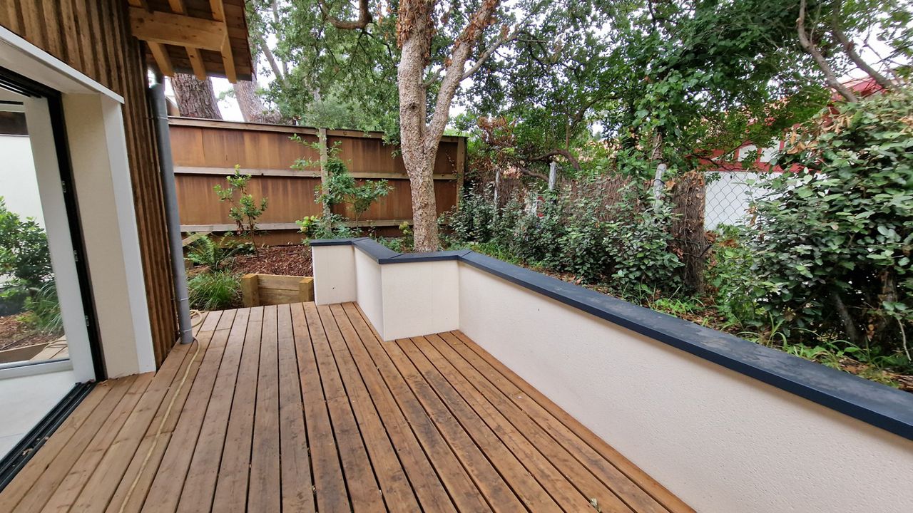 Vue sur une terrasse en bois surélevée avec un muret bas blanc servant de garde-corps et donnant sur un jardin arboré.
